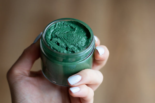 hand holding uncapped glass jar of green spirulina face mask on wooden table