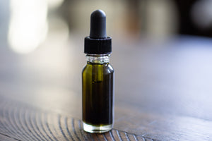A small glass bottle with a dropper top full of dark green liquid sits atop a wooden table